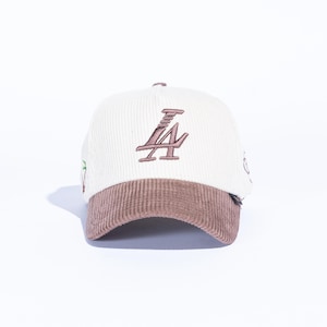 May include: Cream-colored corduroy baseball cap with a brown brim. The cap features the embroidered letters "LA" in brown on the front. A small green and brown design is visible on the side.