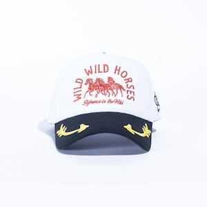 May include: White baseball cap with a black brim. The cap features the words "WILD HORSES" in red, along with a graphic of three horses. The brim has gold-colored decorative accents.