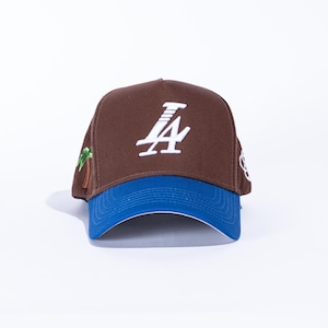 May include: Brown baseball cap with a blue brim. The cap features the white letters "LA" on the front, a palm tree on the side, and a white logo on the other side. The cap is a classic baseball style.