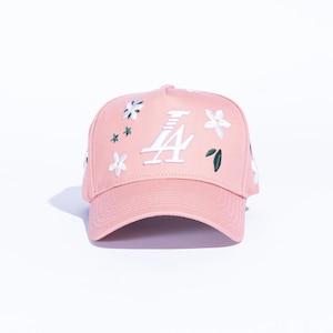 May include: A pink baseball cap with white embroidered flowers and the letter "A" in white. The cap has a curved brim and a structured crown. The floral embroidery includes white flowers and green leaves.
