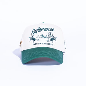 May include: A white baseball cap with a green brim. The cap features the word "Reference" in script above a desert scene with cowboys and cacti. The text "ACE IN THE HOLE" is below the desert scene.
