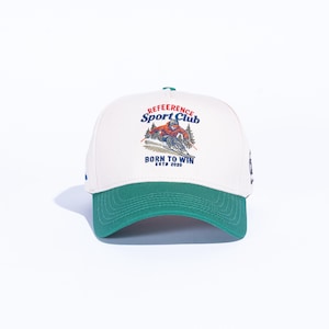 May include: A white baseball cap with a green brim and a graphic of a skier on the front. The cap features the text "REFERENCE Sport Club" and "BORN TO WIN ESTD 2020".