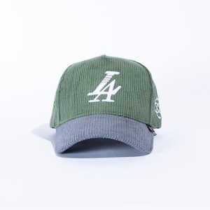 May include: Green corduroy baseball cap with a gray brim. The cap features a white stylized "LA" logo on the front. A small white design is on the side.