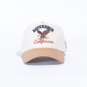 May include: A cream-colored baseball cap with a tan brim. The cap features the word "REFERENCE" above an eagle graphic and the word "California" in red. A red design is on the side of the cap.