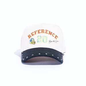 May include: White baseball cap with a black brim and silver studs. The cap features the word "REFERENCE" in orange, the number "20" in blue and yellow, and a football helmet graphic.