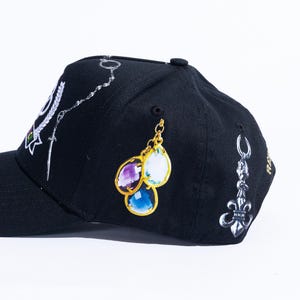 Peut inclure: Une casquette de baseball noire orn&eacute;e d'imprim&eacute;s repr&eacute;sentant une cha&icirc;ne argent&eacute;e avec une &eacute;p&eacute;e, un groupe de trois pierres pr&eacute;cieuses en forme de goutte en violet, blanc et bleu avec des montures dor&eacute;es, et une breloque fleur de lys argent&eacute;e.