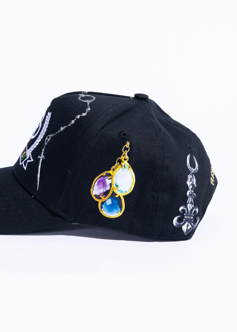 Peut inclure: Une casquette de baseball noire orn&eacute;e d'imprim&eacute;s repr&eacute;sentant une cha&icirc;ne argent&eacute;e avec une &eacute;p&eacute;e, un groupe de trois pierres pr&eacute;cieuses en forme de goutte en violet, blanc et bleu avec des montures dor&eacute;es, et une breloque fleur de lys argent&eacute;e.