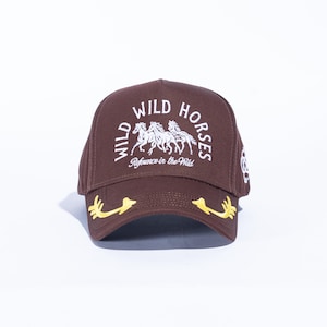 May include: A brown baseball cap with the words "WILD HORSES" in white, along with an image of three horses. The brim features yellow embroidered accents. The text "Refrence in the Wild" is also visible.