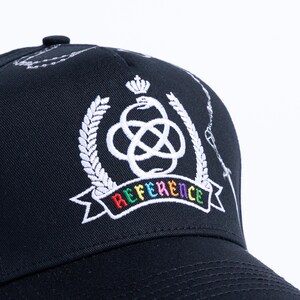 Peut inclure: Une casquette de baseball noire orn&eacute;e d'un embl&egrave;me blanc brod&eacute; repr&eacute;sentant une couronne au-dessus d'un serpent nou&eacute;, entour&eacute; de branches de laurier. Sous l'embl&egrave;me, une banni&egrave;re affiche le mot REFERENCE en lettres brod&eacute;es multicolores.
