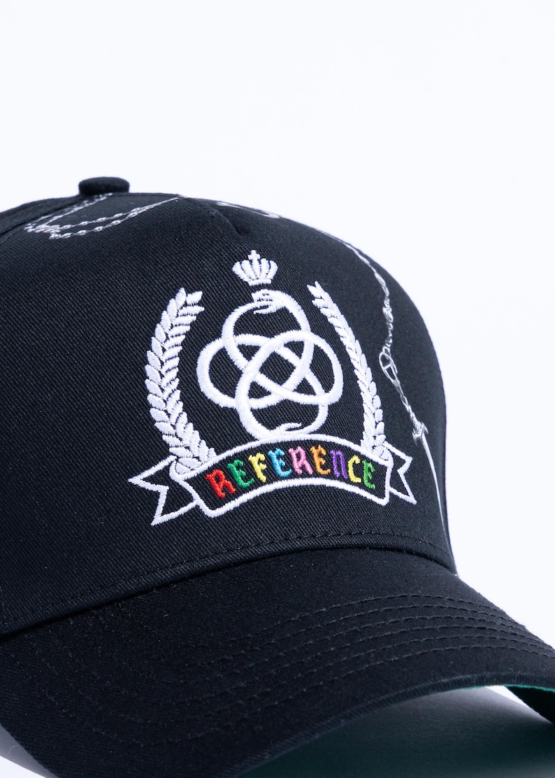 Peut inclure: Une casquette de baseball noire orn&eacute;e d'un embl&egrave;me blanc brod&eacute; repr&eacute;sentant une couronne au-dessus d'un serpent nou&eacute;, entour&eacute; de branches de laurier. Sous l'embl&egrave;me, une banni&egrave;re affiche le mot REFERENCE en lettres brod&eacute;es multicolores.