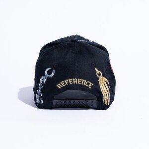 Peut inclure: Une casquette de baseball noire avec une fermeture snapback r&eacute;glable. L'arri&egrave;re de la casquette affiche le mot REFERENCE brod&eacute; en fil dor&eacute;, flanqu&eacute; d'un graphique de breloque m&eacute;tallique argent&eacute;e &agrave; gauche et d'une main squelettique dor&eacute;e.