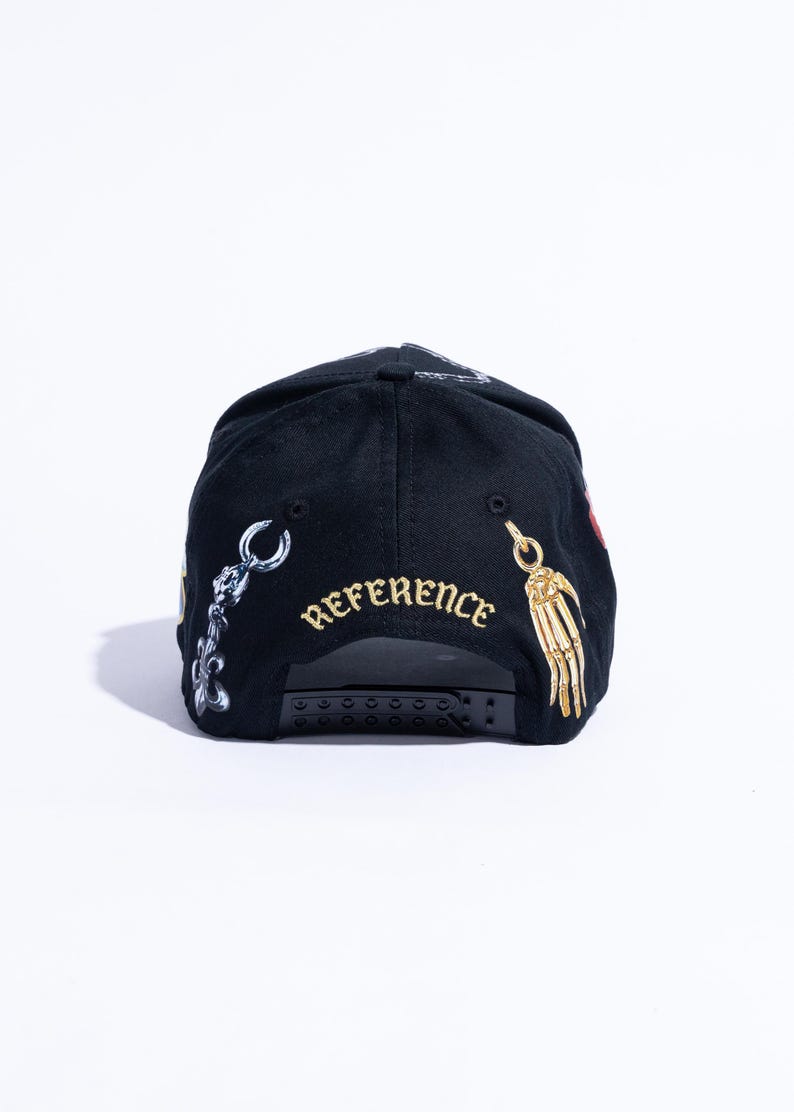 Peut inclure: Une casquette de baseball noire avec une fermeture snapback r&eacute;glable. L'arri&egrave;re de la casquette affiche le mot REFERENCE brod&eacute; en fil dor&eacute;, flanqu&eacute; d'un graphique de breloque m&eacute;tallique argent&eacute;e &agrave; gauche et d'une main squelettique dor&eacute;e.
