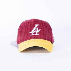 May include: Maroon corduroy baseball cap with a yellow brim. The cap features a white stylized letter "A" on the front. The hat is set against a white background.