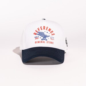 May include: White baseball cap with a navy blue brim. The front of the cap features a red and blue logo with the text "REFERENCE GENERAL STORE" and a flying eagle.