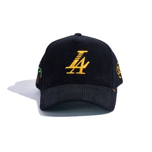 May include: Black corduroy baseball cap with gold embroidered "LA" logo on the front. Features a curved brim and embroidered details on the sides, including a palm tree and a circular design. A stylish accessory.
