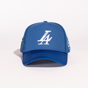 May include: A blue baseball cap with a white "LA" logo embroidered on the front. The cap has a blue mesh back and a blue brim.