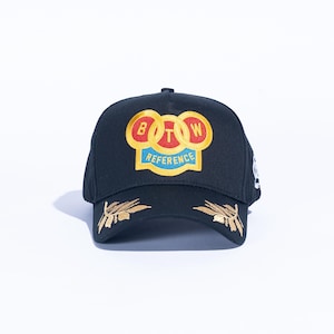 May include: Black baseball cap with gold embroidered leaf accents on the brim. The cap features a colorful patch with the letters "B", "O", and "W" in gold circles, and the word "REFERENCE" in blue.
