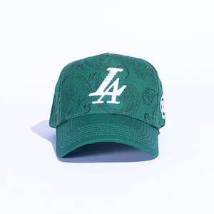 May include: A green baseball cap with a paisley pattern and a white "LA" logo. The cap has a curved brim and a structured crown. The hat is set against a white background.