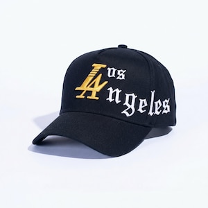 May include: Black baseball cap with the text "LA" in gold and "Angeles" in white, embroidered on the front. The cap has a curved brim and a button on top. The hat is made of a durable fabric.