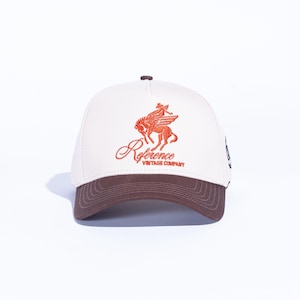 May include: A baseball cap with a cream-colored crown and a brown brim. The cap features an orange graphic of a winged horse and rider, with the word "Reference" in cursive and "Vintage Company" below.