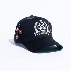 Peut inclure: Une casquette de baseball noire orn&eacute;e d'un embl&egrave;me blanc brod&eacute; repr&eacute;sentant un serpent enroul&eacute; autour d'un n&oelig;ud, surmont&eacute; d'une couronne et entour&eacute; de branches de laurier. Sous l'embl&egrave;me, une banni&egrave;re affiche le mot REFERENCE en lettres multicolores.