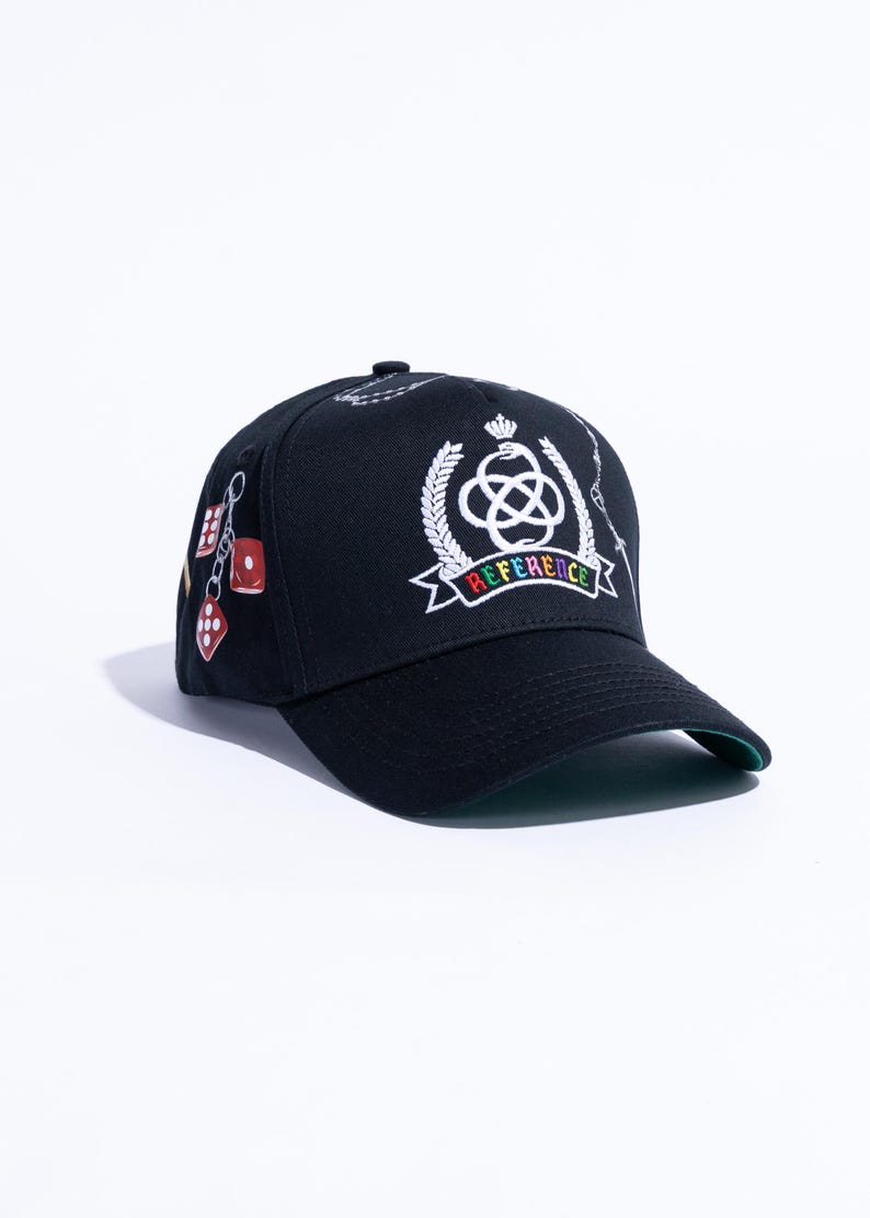 Peut inclure: Une casquette de baseball noire orn&eacute;e d'un embl&egrave;me blanc brod&eacute; repr&eacute;sentant un serpent enroul&eacute; autour d'un n&oelig;ud, surmont&eacute; d'une couronne et entour&eacute; de branches de laurier. Sous l'embl&egrave;me, une banni&egrave;re affiche le mot REFERENCE en lettres multicolores.