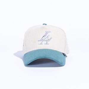 May include: Cream-colored corduroy baseball cap with a teal brim. The cap features the embroidered letters "LA" in a silver color on the front. The hat is set against a white background.