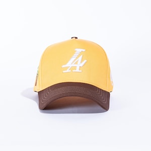 May include: A yellow baseball cap with a brown brim. The cap features a white embroidered "A" logo on the front. The cap is a classic six-panel design, suitable for casual wear.