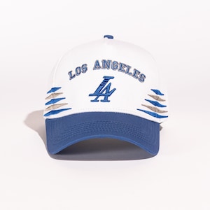 May include: White baseball cap with a blue brim and a blue and silver Los Angeles Dodgers logo. The cap has a white mesh back and a silver "Los Angeles" text.