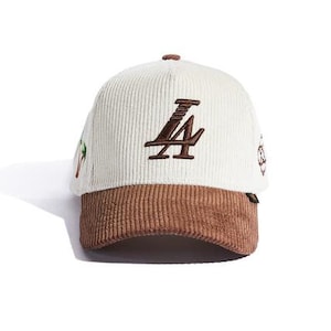 May include: A corduroy baseball cap with a white crown and a brown brim. The cap features the embroidered letters "LA" in brown on the front. Small embroidered details are visible on the sides.