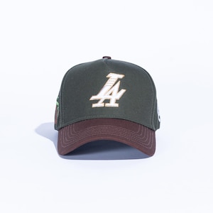May include: A dark green baseball cap with a brown brim. The cap features a white and gold embroidered "LA" logo on the front. The cap is a classic style, suitable for casual wear.