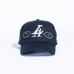 May include: Black baseball cap with white "LA" logo and rhinestone accents. The cap features a curved brim and a structured crown. The design includes decorative elements around the logo, and a small design on the side.