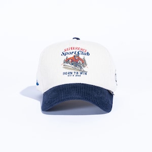 May include: A white corduroy baseball cap with a navy blue brim. The cap features an embroidered graphic of a skier and the text "REFERENCE Sport Club" and "BORN TO WIN ESTD 2020".