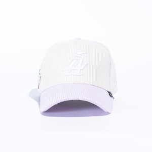 May include: Cream-colored corduroy baseball cap with a lavender brim. The cap features a white embroidered logo on the front. A small design is visible on the side.