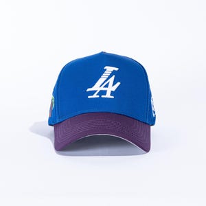 May include: A blue baseball cap with a purple brim. The cap features a white logo on the front and small embroidered details on the sides. The cap is made of a durable fabric and has a curved brim.