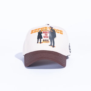 May include: Cream-colored baseball cap with a brown brim. The cap features a graphic of two figures and the text "REFERENCE" and "BORN TO WIN" in orange. A black design is on the side.