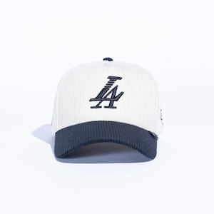 May include: Cream-colored corduroy baseball cap with a navy blue brim. The cap features the embroidered letters "LA" in navy blue on the front. The hat has a curved brim and a small navy blue button on top.