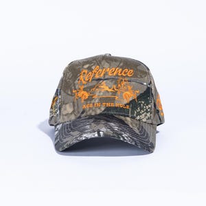 May include: Camouflage baseball cap with the word "Reference" embroidered in orange across the front. Below the word "Reference" is an orange graphic and the text "ACE IN THE HOLE". The cap has a curved brim and a structured crown.