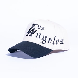 May include: Cream-colored baseball cap with a navy blue brim. The cap features the embroidered text "LA Los Angeles" in a gothic font. A stylish accessory for casual wear.