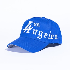 May include: A royal blue baseball cap with the text "Los Angeles" in white gothic lettering. The cap has a curved brim and a structured crown. The hat is set against a white background.