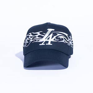 May include: Black baseball cap with white embroidered "LA" logo and flame design. The cap has a curved brim and is set against a white background. A stylish accessory for casual wear.