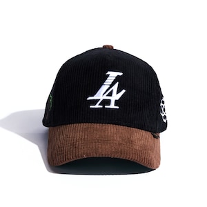 May include: Black corduroy baseball cap with a brown brim. The cap features white embroidered text "LA" on the front. Additional embroidered details are visible on the side. The cap is set against a white background.
