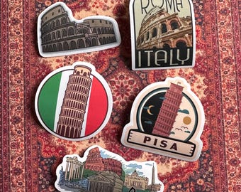 May include: Five colorful stickers featuring iconic landmarks of Rome and Pisa, Italy. The stickers feature illustrations of the Colosseum, the Trevi Fountain, the Leaning Tower of Pisa, and the Pantheon. The stickers are printed on white paper with black outlines and have a glossy finish.