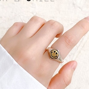 May include: A silver ring with a gold smiley face design. The ring has a geometric shape and is worn on a finger. The text "PICK • A • SMILE" is visible at the top of the image.