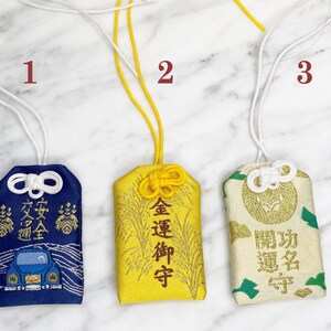 May include: Three Japanese omamori charms with white cords. The first is navy blue with a car graphic and Japanese text. The second is gold with Japanese text. The third is cream with green accents and Japanese text.