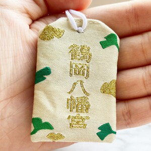 May include: A rectangular, cream-colored Japanese omamori charm with gold and green accents. The charm features gold text and green leaf-like shapes. A white string is attached at the top.