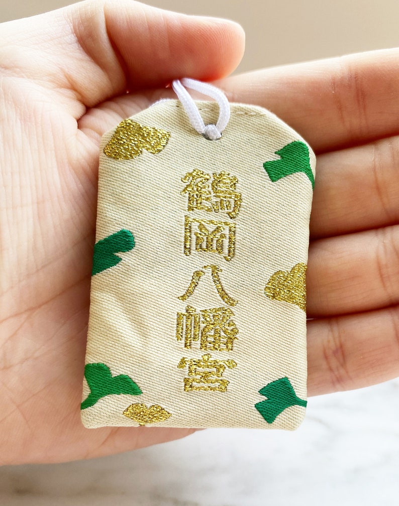 May include: A rectangular, cream-colored Japanese omamori charm with gold and green accents. The charm features gold text and green leaf-like shapes. A white string is attached at the top.