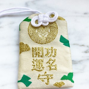 May include: Cream-colored Japanese omamori charm with gold embroidery and green accents. The charm features Japanese characters and a decorative knot. A white cord is attached for hanging.