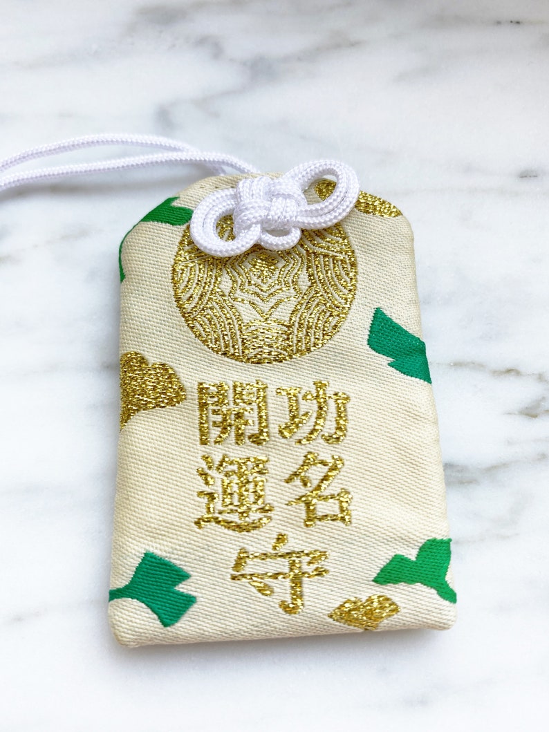 May include: Cream-colored Japanese omamori charm with gold embroidery and green accents. The charm features Japanese characters and a decorative knot. A white cord is attached for hanging.
