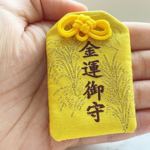May include: A yellow Japanese omamori charm with gold accents and kanji characters. The charm has a decorative knot at the top and a gold embroidered design. The text on the charm is in dark brown.